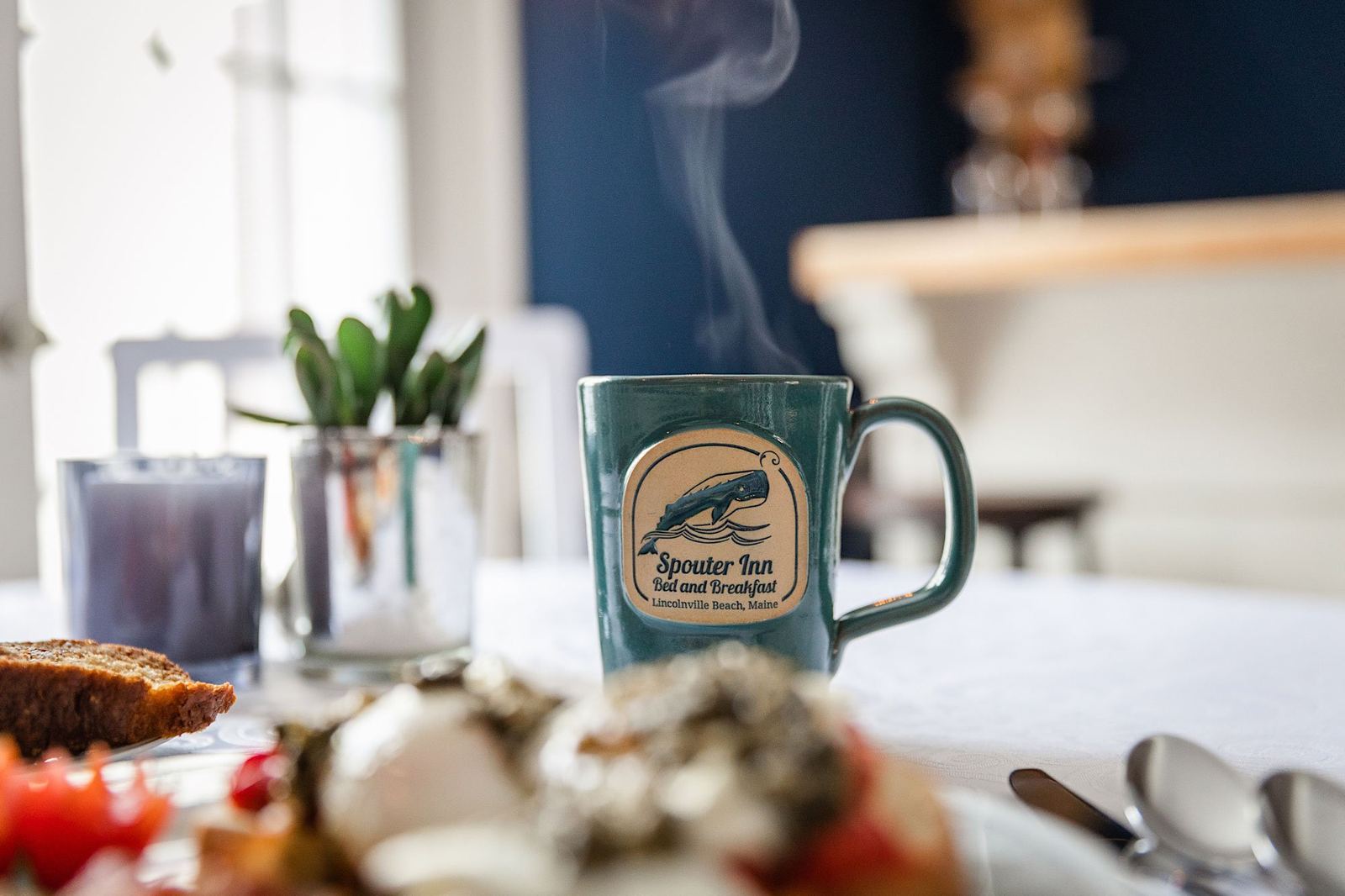 A steaming dark-green Spouter Inn whale-logo mug next to a breakfast plate
