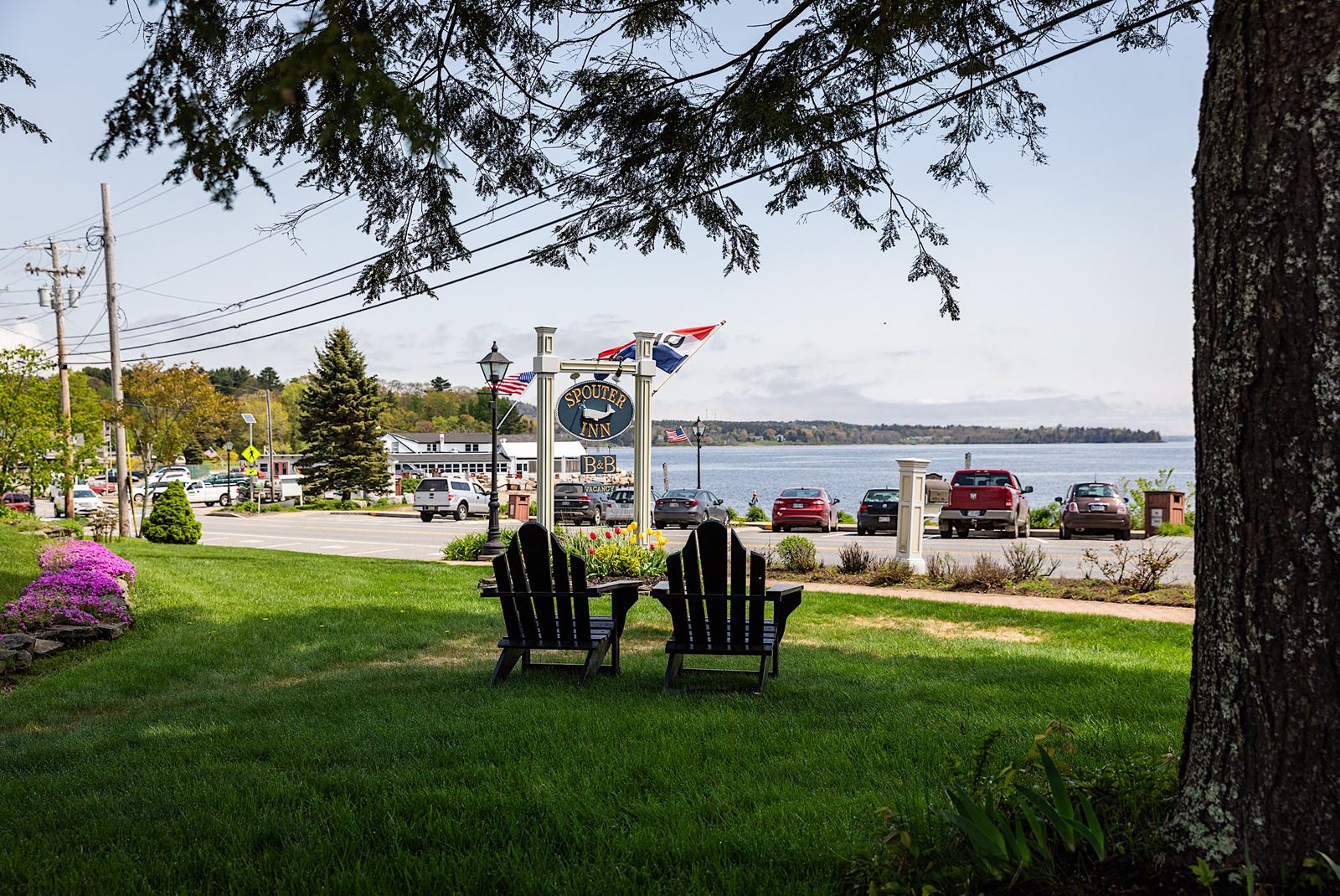 Two Adirondack chairs on the inn's front lawn with the Spouter Inn sign, flags, and Penobscot Bay beyond